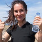 Happy woman giving thumbs up with water bottle on a sunny beach day in Portugal.
