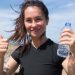 Happy woman giving thumbs up with water bottle on a sunny beach day in Portugal.